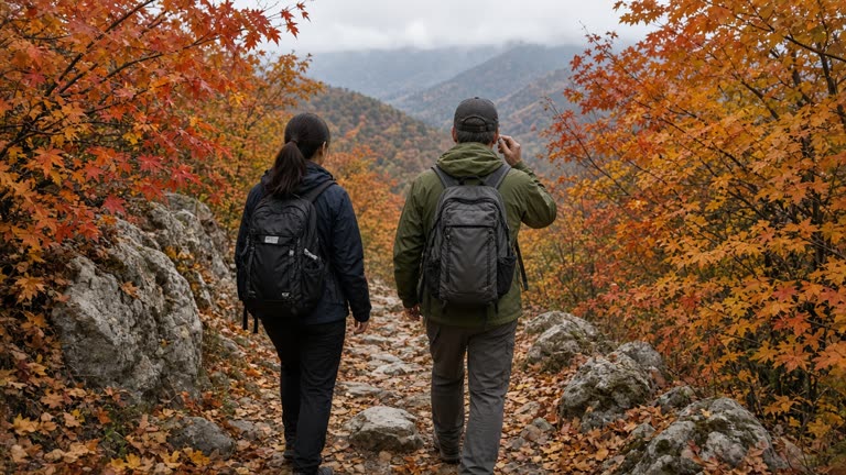 Hikers on a rocky autumn mountain trail seen from behind, one applying a small sunscreen stick, maple foliage and overcast daylight, backpacks without readable brand logos.