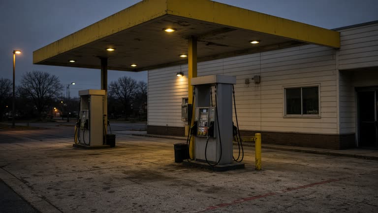 A corner convenience store fuel island with yellow canopy and worn concrete forecourt at dusk under sodium vapor lights, pump faces turned away from camera, no logos or readable text