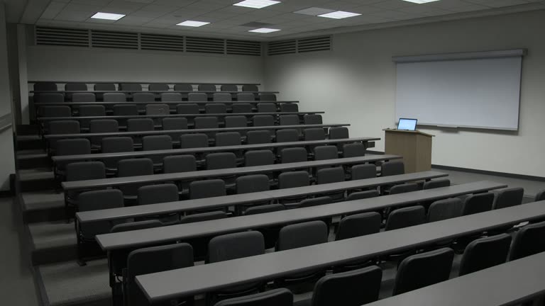 Tiered empty university lecture hall seats facing a blank projection screen under cool fluorescent light, a lone laptop on the lectern with screen glow but no readable text