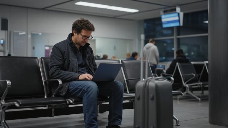 Traveler seated at an airport gate with a laptop open on their knees under cool terminal lighting, screen glow without readable text, no logos