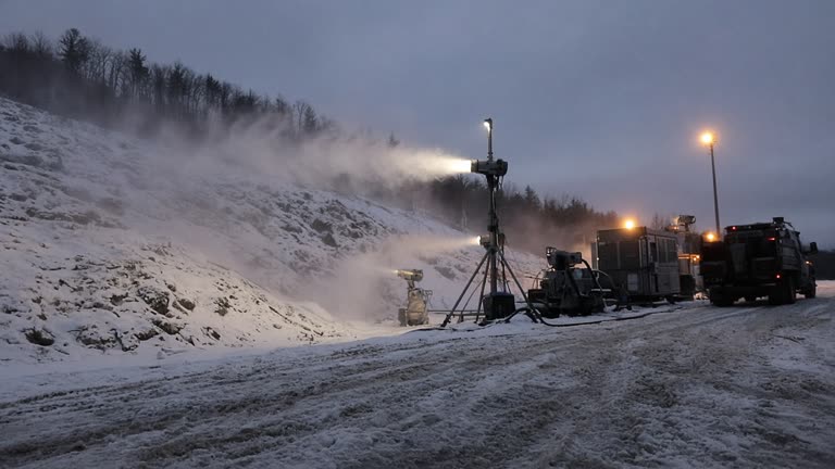 Ski resort snowmaking guns spraying mist on a bare slope at dusk with compressor equipment and sodium yard lights, packed snow and service vehicles, no logos or readable text