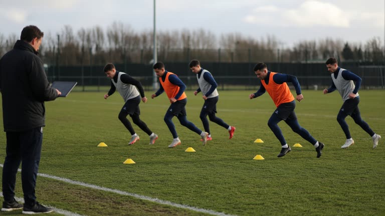 Professional footballers in training bibs performing cone drills on a grass pitch, sports scientist with tablet on sideline, afternoon sun, no logos or readable text.