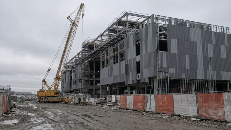 Semiconductor fab construction site at overcast dawn with crane beside a partially clad building, mud-tracked service road, safety fencing, no logos or readable text