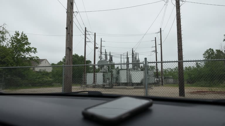 Wooden utility poles and a small electrical substation behind a chain-link fence in humid Atlanta suburbs, overcast afternoon, a smartphone lying on a car dashboard in soft focus foreground, no logos or readable text