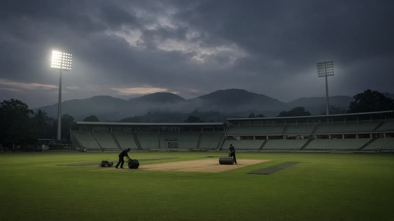 Evening view of a cricket stadium outfield under floodlights with empty green seats and groundstaff rolling the pitch, Jharkhand hills in haze, no logos or readable text.
