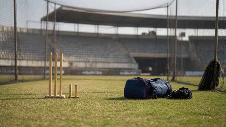 Cricket practice nets with empty stumps and kit bags on grass, afternoon sun, stadium stands blurred in background, no logos or readable text