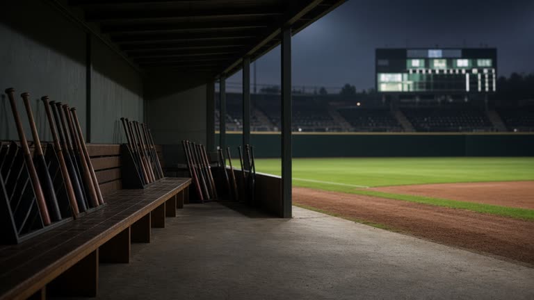 Empty baseball dugout with bats in racks and a distant outfield scoreboard glowing, evening stadium lights, no logos or readable text